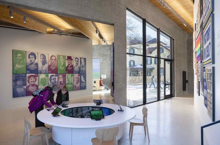 Photo of the interior of the Equal Rights Heritage Center which shows two people interacting with a digital exhibit on an iPad. Behind them is a wall of portraits of key figures in U.S. history