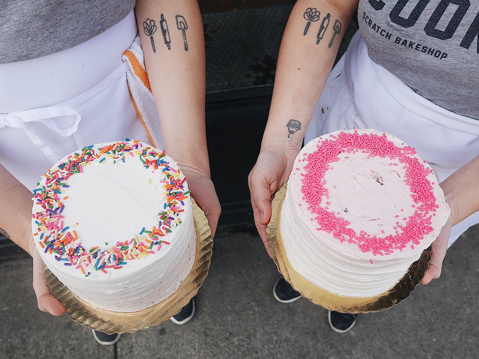 Photo of two people holding frosted cakes. One is white with rainbow sprinkles and the other is white with pink sprinkles