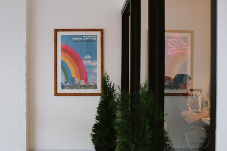 Photo of the interior of TGW offices with a green tree, office windows, and a poster that has a rainbow over Rochester with the text 'Rochester Monroe County, NY, a great place to do business'