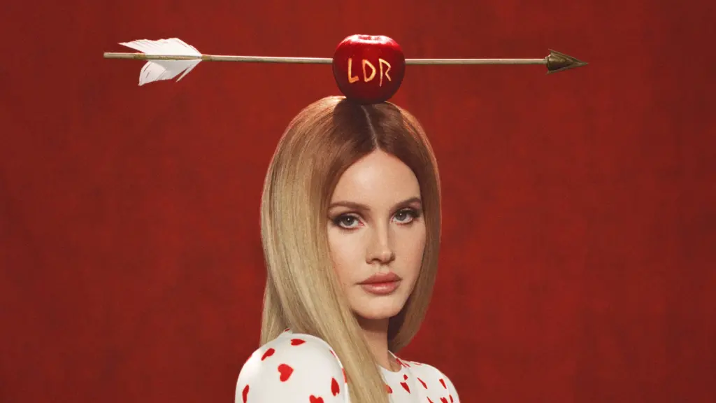 Lana Del Rey poses against a red background with a red apple on top of her head with the initials "LDR" carved in it with an arrow through it.