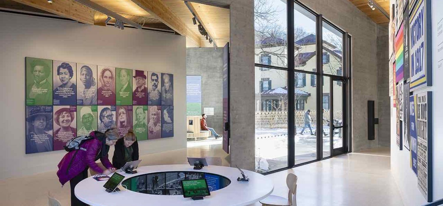 Photo of the interior of the Equal Rights Heritage Center which shows two people interacting with a digital exhibit on an iPad. Behind them is a wall of portraits of key figures in U.S. history
