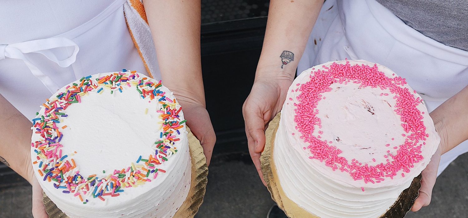 Photo of two people holding frosted cakes. One is white with rainbow sprinkles and the other is white with pink sprinkles