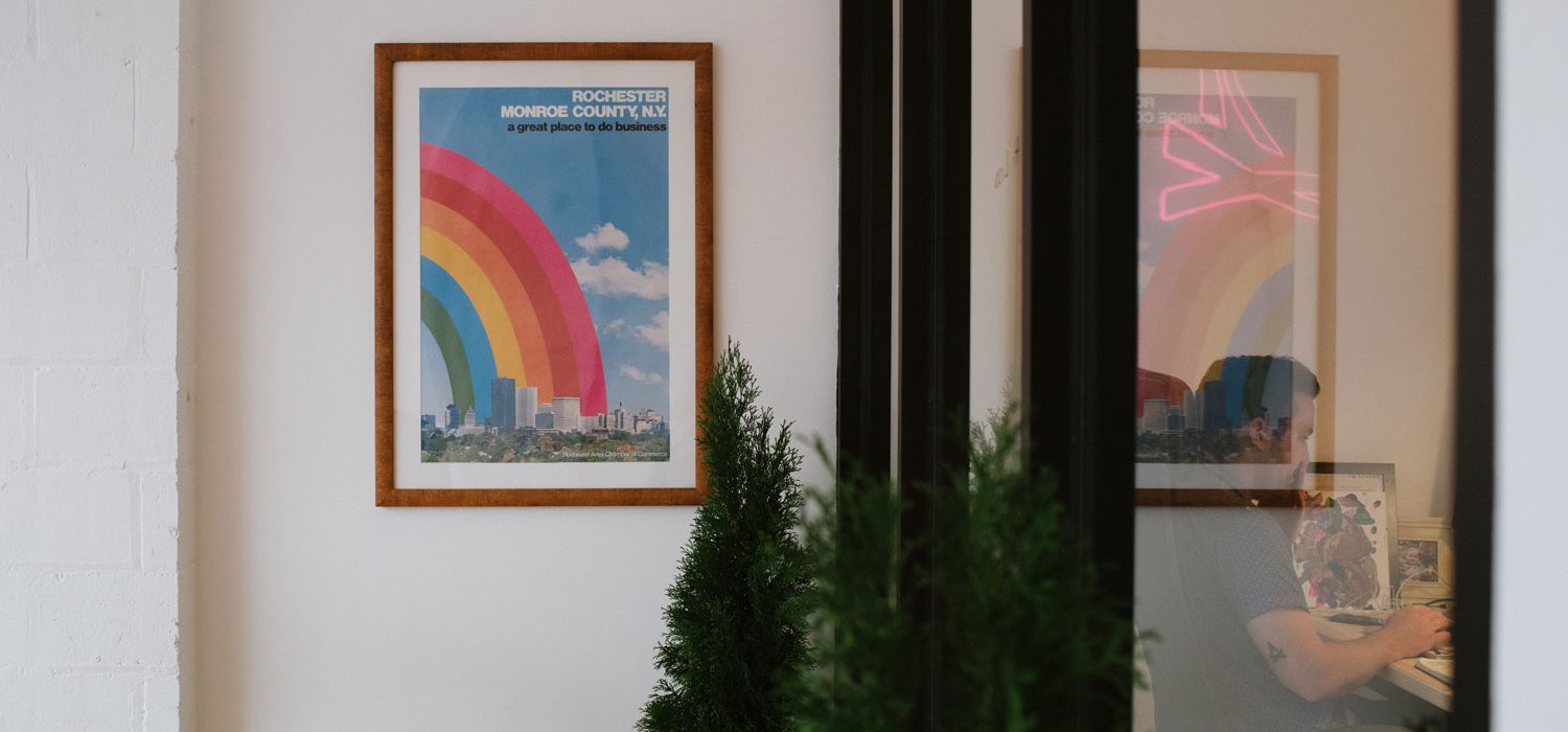 Photo of the interior of TGW offices with a green tree, office windows, and a poster that has a rainbow over Rochester with the text 'Rochester Monroe County, NY, a great place to do business'